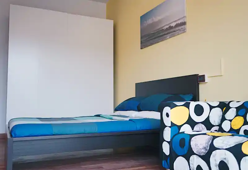 Dormitorio moderno y luminoso con cama doble con ropa de cama azul, armario blanco y un sillón estampado. Líneas limpias y colores tranquilos de inspiración costera.