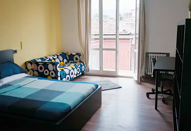 Dormitorio moderno y luminoso con cama doble, sofá estampado y grandes puertas al balcón que llenan la habitación de luz natural.