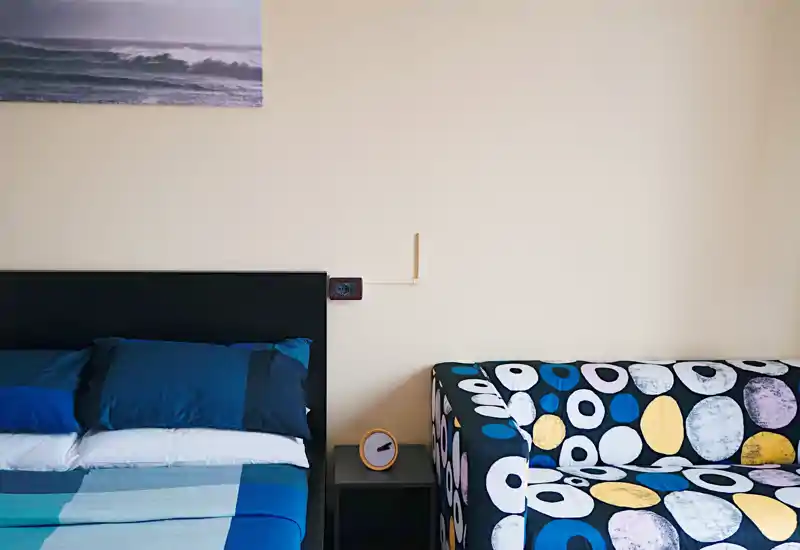 Rincón de dormitorio luminoso y moderno con una cama doble bien hecha y un sofá estampado y colorido, ideal para estudios o habitación de invitados.