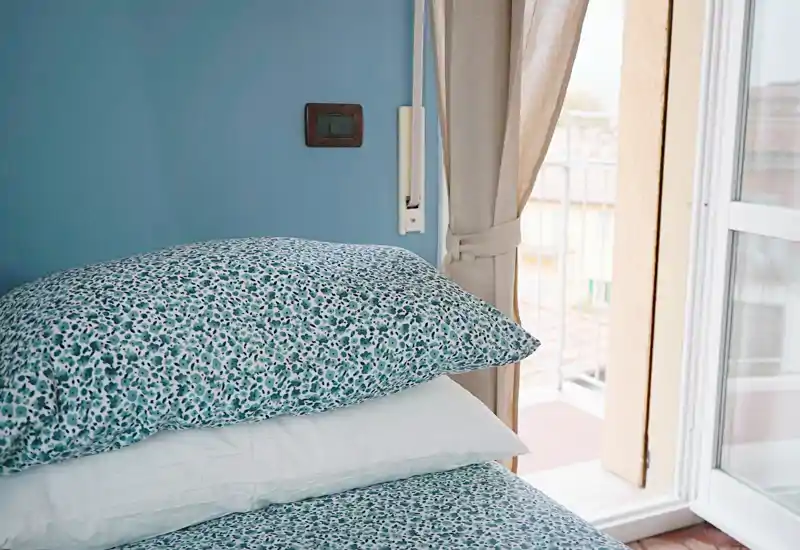 Rincón de dormitorio luminoso y acogedor con cojines estampados y luz natural desde el balcón; ropa de cama sencilla y pared azul para una atmósfera relajante.