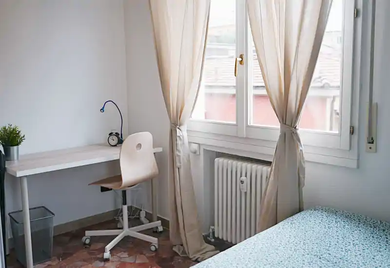Rincón de dormitorio luminoso y minimalista con una cama individual parcialmente visible, un pequeño escritorio y una ventana con cortinas ligeras que dejan pasar la luz natural.