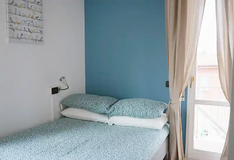Dormitorio ordenado y luminoso con cama doble vestida con ropa de cama estampada, pared azul como acento y ventana al balcón que aporta luz natural.