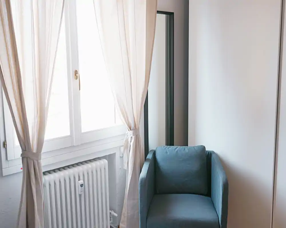 Rincón de salón luminoso y minimalista con un sillón cómodo junto a la ventana con cortinas claras — ideal como rincón de lectura.