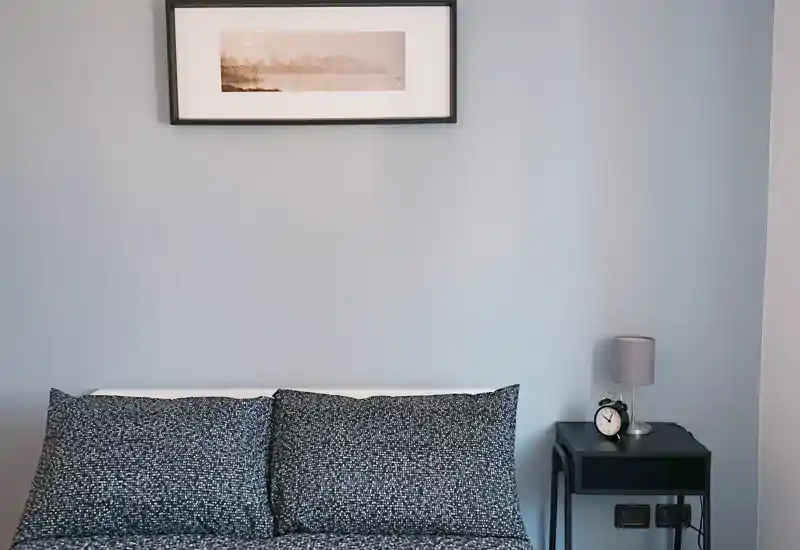 Imagen de dormitorio bien presentada con cama doble y ropa de cama estampada, mesita con lámpara y despertador y un cuadro encima: tonos neutros y aspecto ordenado.