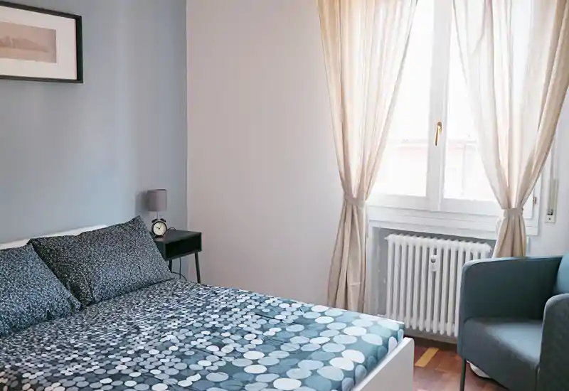Camera da letto luminosa e ben curata con biancheria a fantasia, comodino e poltrona accanto a una grande finestra che illumina dolcemente l'ambiente.