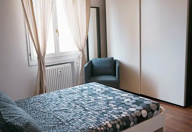 Dormitorio luminoso y sencillo con una cama doble con funda estampada, un sillón junto a la ventana y un amplio armario. La luz natural y los tonos neutros crean un ambiente tranquilo, ideal para el anuncio.