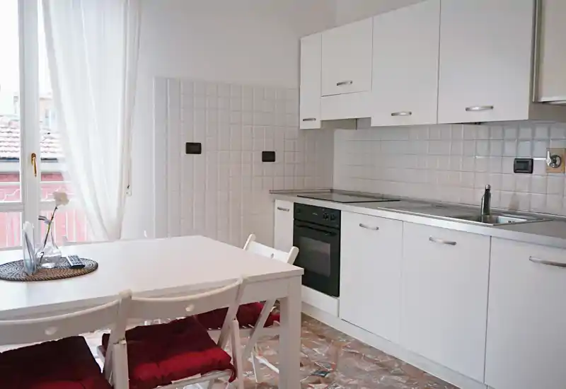 Cucina moderna e luminosa con mobili bianchi e piccolo tavolo da pranzo vicino a una finestra ampia, con sedie imbottite e pavimento effetto marmo.