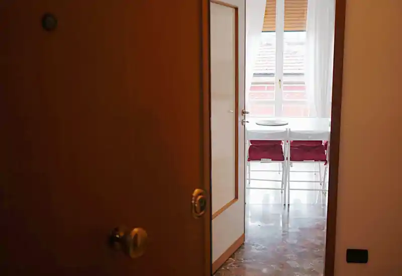 View from an open front door into the apartment hallway leading to a bright area with a small table and chairs; polished floor and natural light from the window beyond.