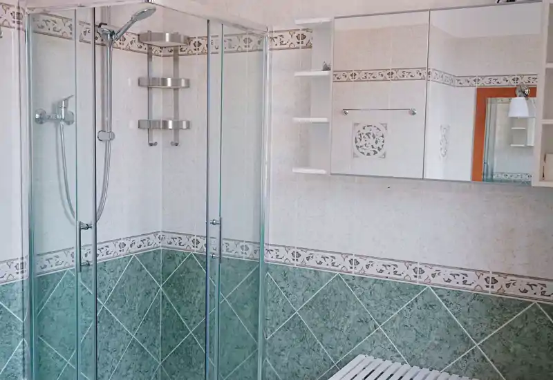 Clean, tiled bathroom with a glass corner shower, wall-mounted mirrored cabinet and decorative green lower tiles.