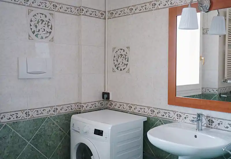 Compact bathroom with tiled walls, a pedestal sink, mirror and a washing machine — bright and well-kept, suitable for rental listings.