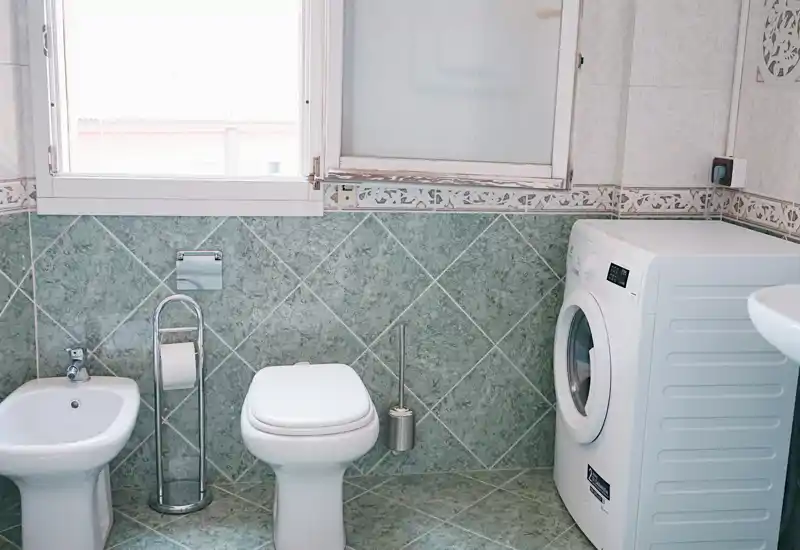 Bathroom with bidet, toilet and a washing machine; green tiled walls and floor, bright natural light from a frosted window.