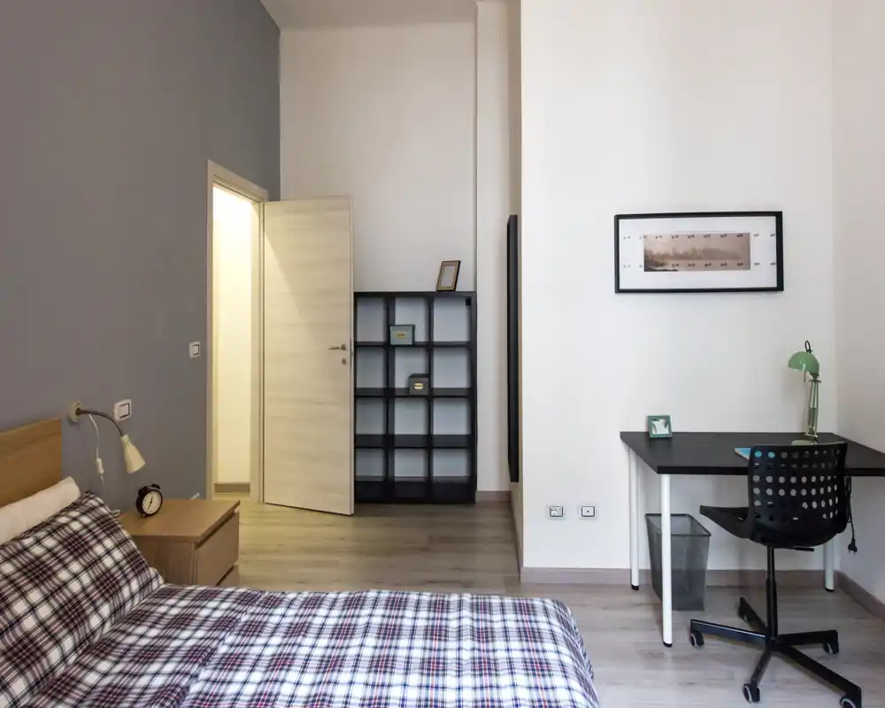 Bright, modern bedroom with a double bed dressed in patterned bedding, a bedside table and lamp, shelving unit and a study desk with chair — calm, functional layout ideal for a rental listing.