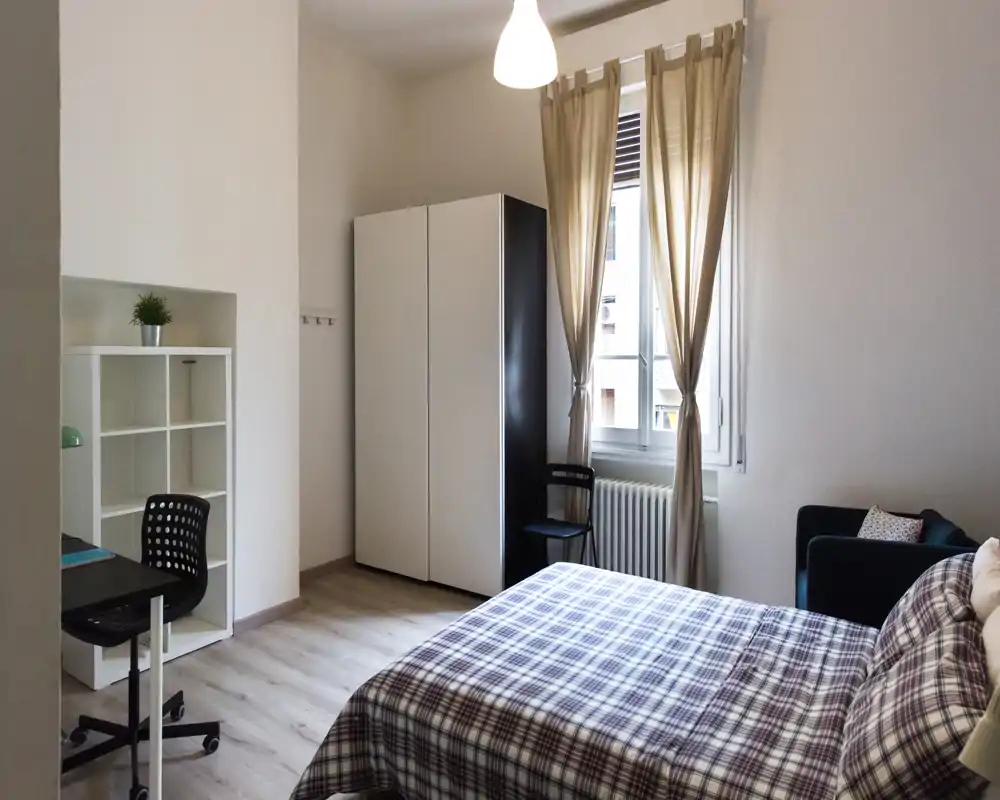 Bright, well-staged bedroom with a double bed in checked linen, wardrobe, small desk and armchair beside a large window letting in natural light.
