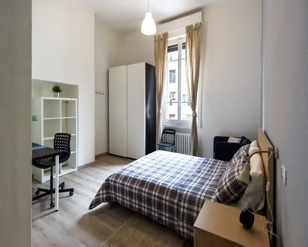 Bright, well-styled bedroom with a double bed in plaid bedding, wardrobe, desk and a small seating area by the window — ideal for a comfortable student or single professional stay.