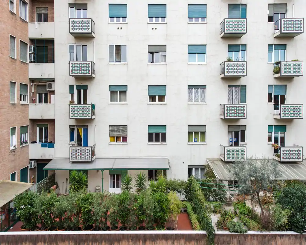 Urban courtyard and small garden area in front of a multi-storey apartment façade, with potted plants and several small balconies.