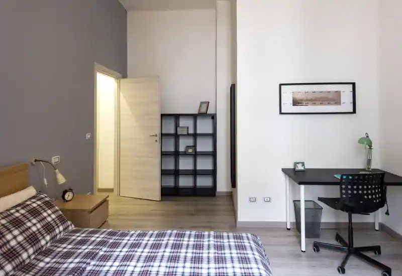 Bright, modern bedroom with a double bed dressed in patterned bedding, a bedside table and lamp, shelving unit and a study desk with chair — calm, functional layout ideal for a rental listing.