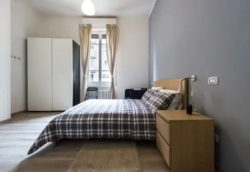 Bright, modern bedroom with a double bed dressed in checkered bedding, a wooden bedside table with lamp, wardrobe and natural light from a window.