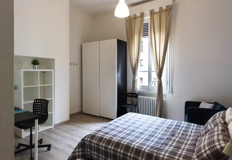 Bright, well-staged bedroom with a double bed in checked linen, wardrobe, small desk and armchair beside a large window letting in natural light.