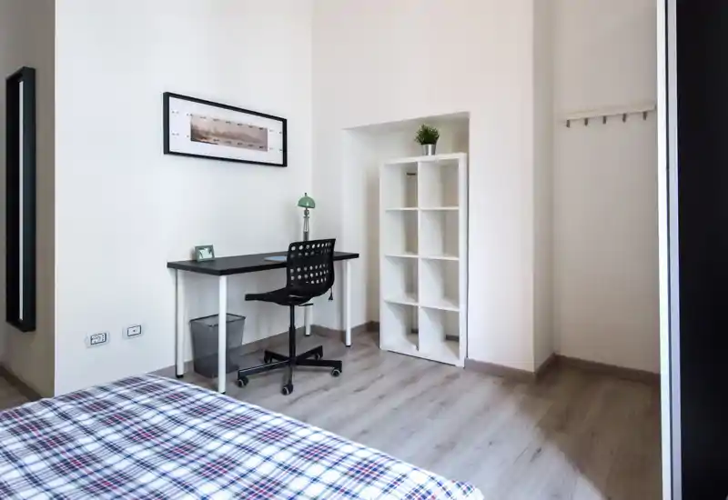 A bright, minimalist bedroom corner with a partially visible bed in the foreground, a work desk with chair and lamp, and a white shelving unit—clean styling and natural light.