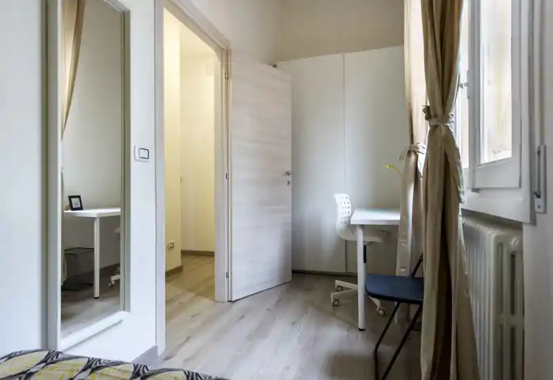 Rincón de dormitorio pequeño y ordenado con luz natural, escritorio compacto, armario y parte de la cama visible en primer plano.