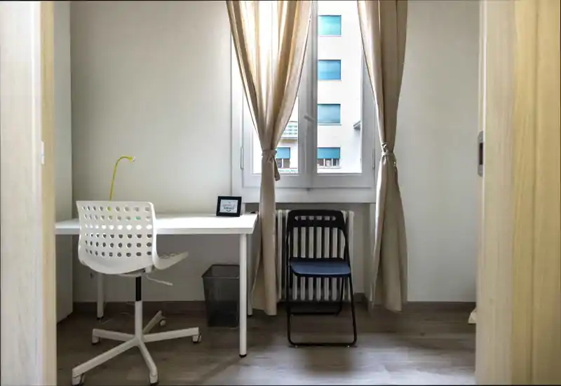 Rincón de dormitorio luminoso y minimalista con un escritorio sencillo, dos sillas y una ventana con cortinas neutras; ideal como zona de estudio en la habitación.