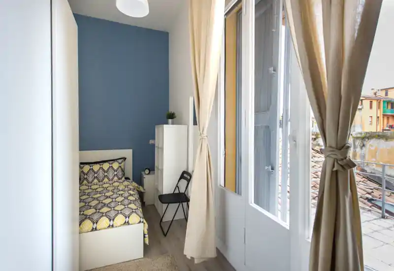 Chambre compacte et lumineuse avec un lit simple aux draps à motifs, située à côté d'une grande fenêtre aux rideaux clairs, avec une petite armoire et une chaise pliante.