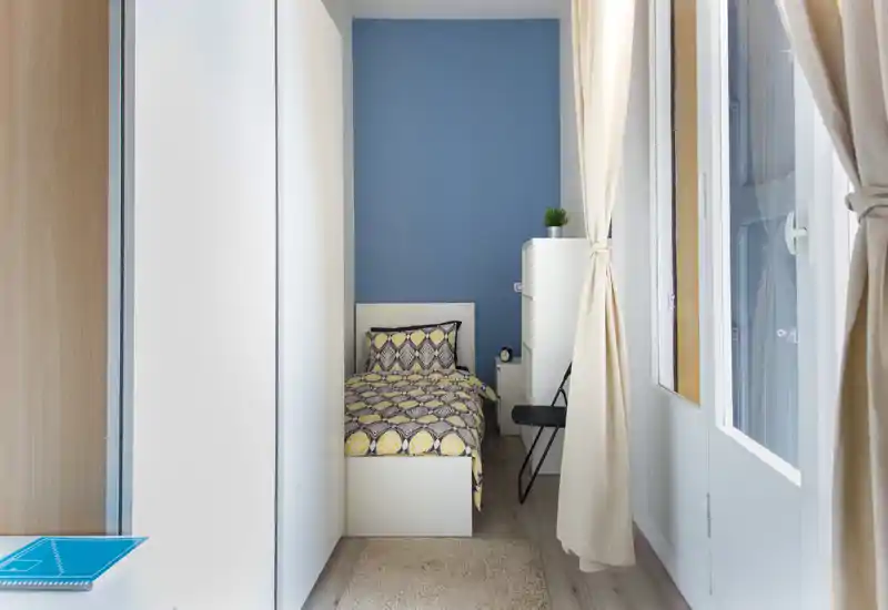Petite chambre simple avec une couette à motif, un sol clair et une grande fenêtre apportant une belle lumière naturelle. Mobilier sobre avec une petite armoire, un tapis et une chaise — idéale pour une location lumineuse et confortable.