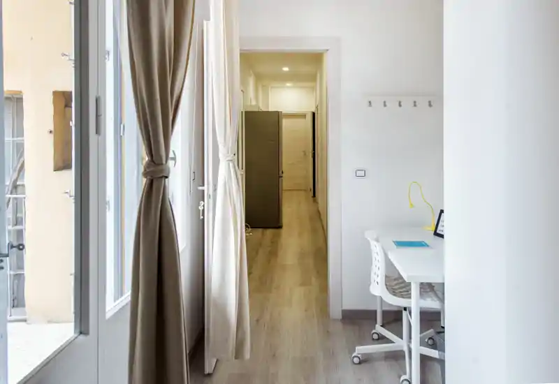 Entrée/couloir lumineux avec un petit coin bureau : lumière naturelle par de grandes fenêtres, sol en bois clair et un petit bureau blanc avec chaise — idéal pour un studio où l'entrée sert aussi d'espace de travail.