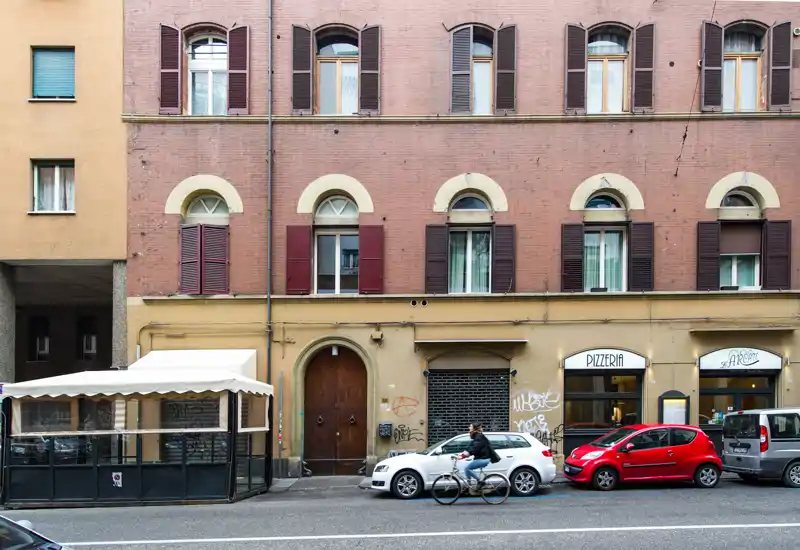 Facciata a livello strada con ingresso di edificio, una pizzeria, auto parcheggiate e un dehors coperto; scatto esterno urbano chiaro, adatto per descrivere la parte commerciale/residenziale al piano terra.