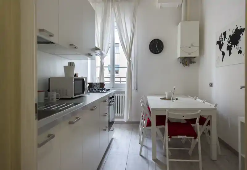Cucina moderna e luminosa con mobili bianchi, piano cottura a gas e tavolo da pranzo per quattro. La luce naturale da una finestra alta e l’arredo semplice creano un ambiente pulito e accogliente.