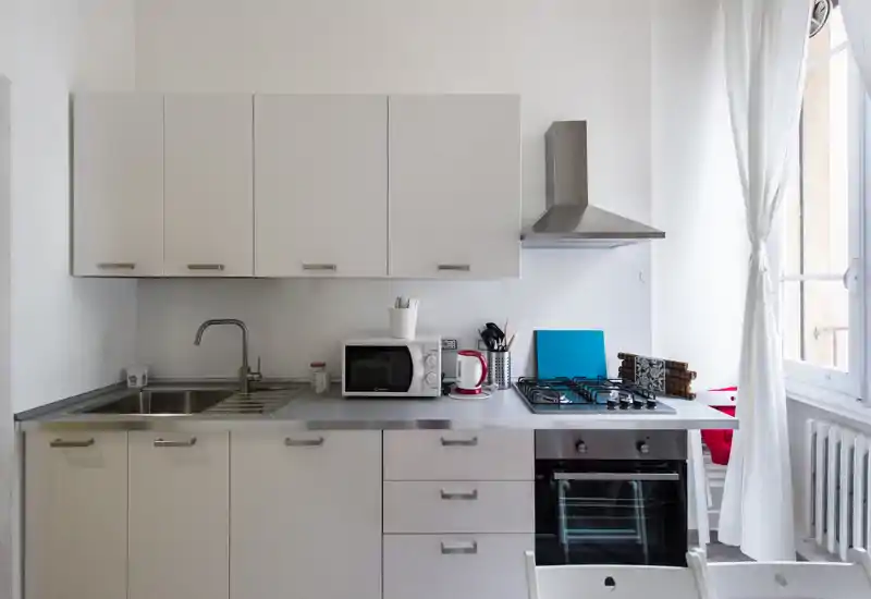 Cucina moderna e luminosa con pensili bianchi, lavello in acciaio, piano cottura a gas e forno incassato; la luce naturale dalla finestra rende l'ambiente arioso e pulito, perfetta per monolocali.