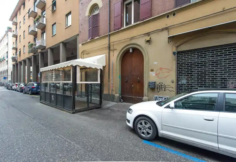 Scena urbana prospiciente la strada con facciate di edifici, un'auto parcheggiata e una struttura coperta sul marciapiede; adatta a mostrare l'ingresso esterno e il parcheggio stradale vicino.