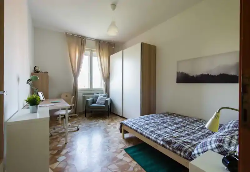Camera singola ben illuminata con letto comodo, scrivania per lo studio, poltrona e ampia armadiatura. Toni neutri e luce naturale rendono lo spazio tranquillo e funzionale.
