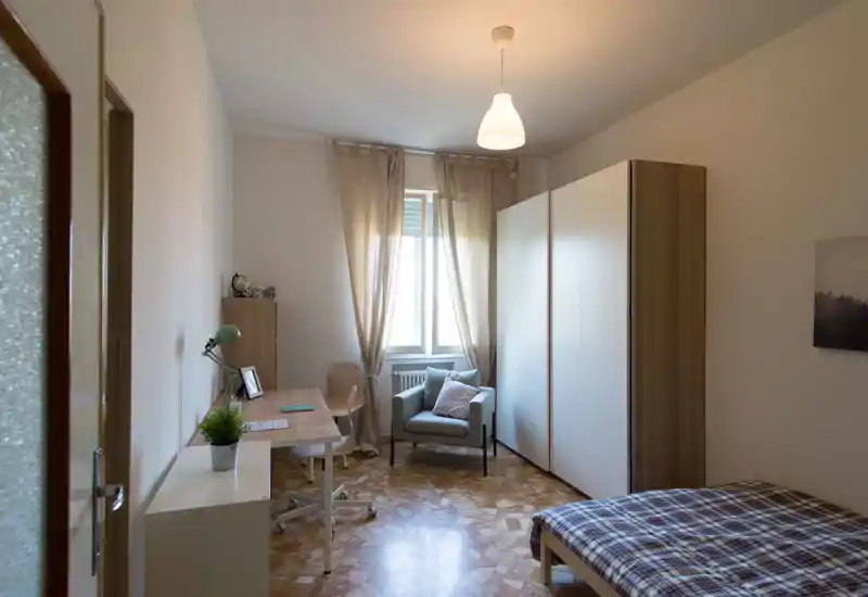 Camera da letto luminosa e arredata con semplicità: letto singolo, armadio, scrivania e poltrona vicino alla finestra; tende neutre e luce naturale creano un'atmosfera calma e accogliente.