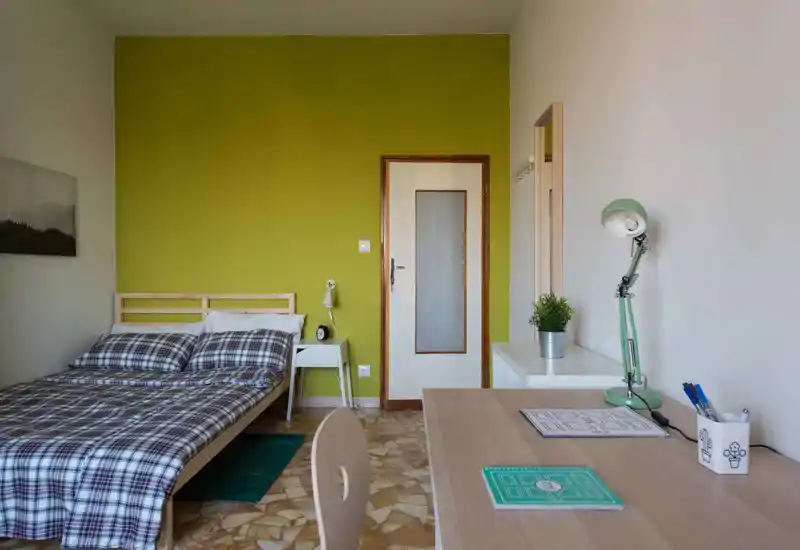 Camera da letto luminosa e ordinata con letto matrimoniale, comodino e scrivania. Parete verde a contrasto e arredi essenziali creano un’atmosfera fresca e accogliente, perfetta per un annuncio.