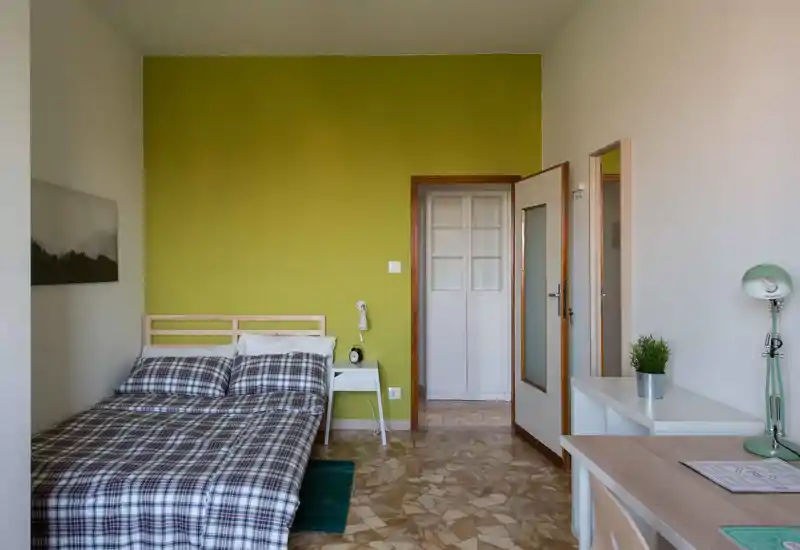 Camera da letto luminosa e ordinata con letto matrimoniale a fantasia davanti a una parete verde, comodino e angolo scrivania — perfetta per soggiorni comodi e lavoro in stanza.