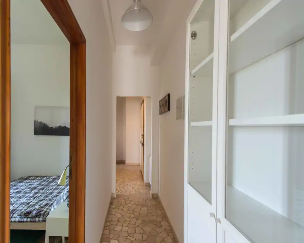 Corridoio luminoso e stretto con pavimento a motivo in pietra, pareti bianche e vista parziale di una camera a sinistra; ingresso semplice e pulito adatto agli annunci di appartamento.