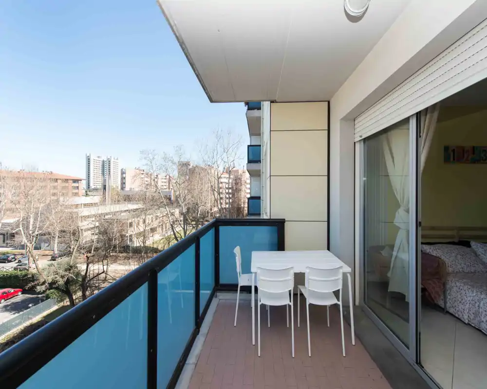 Balcone soleggiato con tavolo e quattro sedie, parapetto a pannelli di vetro e vista sul quartiere — ideale per pranzi all'aperto o relax.