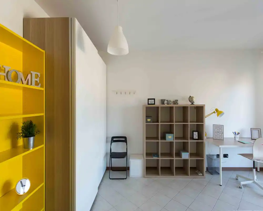 Zona giorno luminosa e minimale con scaffalatura aperta, angolo scrivania e tocchi di giallo. Lo spazio è pulito e semplicemente arredato, adatto per lavorare e conservare oggetti.