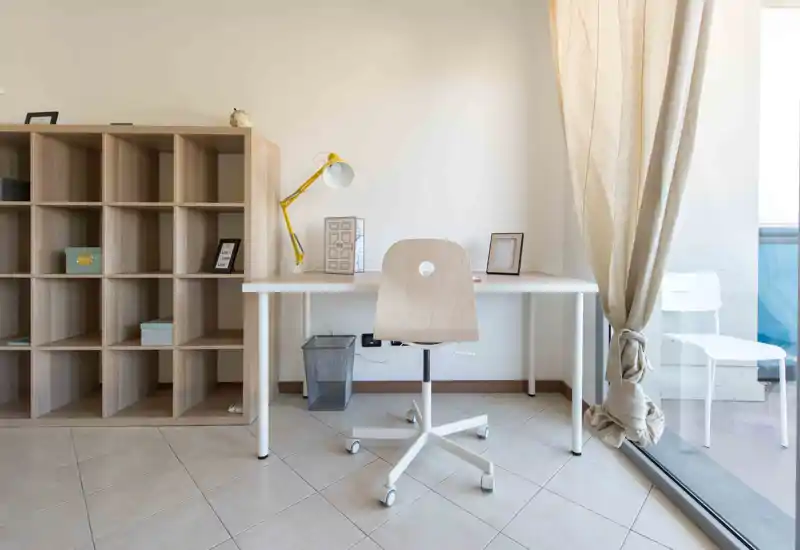 Zona giorno luminosa e minimal con scrivania ordinata e scaffalatura — un angolo studio tranquillo vicino a una grande finestra con luce naturale e arredi neutri.