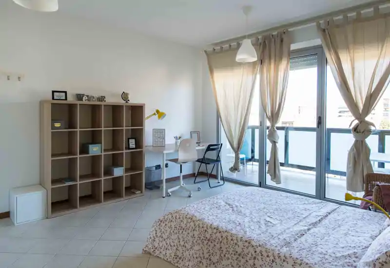 Camera da letto luminosa e ariosa con letto matrimoniale in primo piano, libreria modulare e una piccola scrivania vicino alle porte scorrevoli che danno sul balcone. Toni neutri e tende morbide creano un’atmosfera calma e accogliente, perfetta per annunci di affitto.