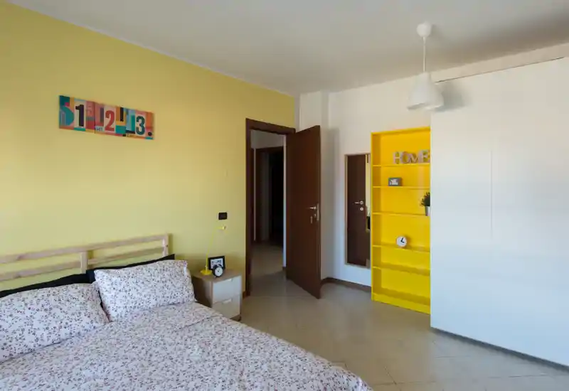 Camera da letto luminosa e ordinata con letto matrimoniale, comodino con lampada e una vivace scaffalatura gialla come elemento decorativo.