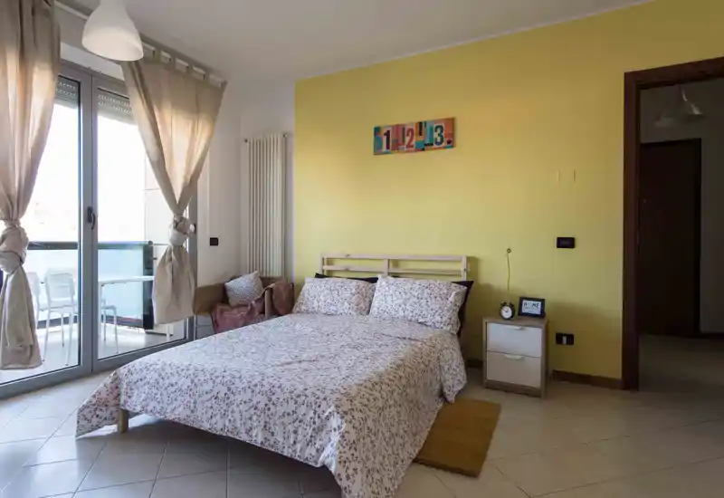 Camera da letto luminosa e ben arredata con letto matrimoniale, biancheria a fantasia, comodino e accesso al balcone con luce naturale.