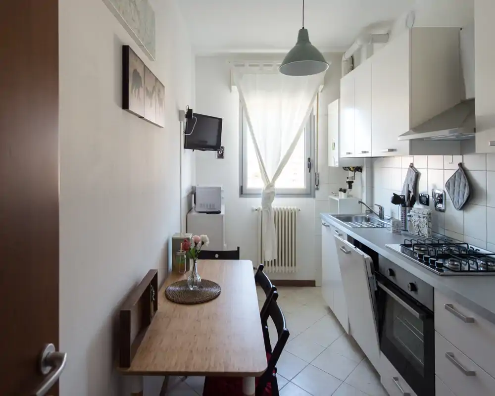 Cucina compatta e luminosa con piano cottura a gas, forno e un piccolo tavolo da pranzo vicino alla finestra; ordinata, moderna e ben illuminata dalla luce naturale.