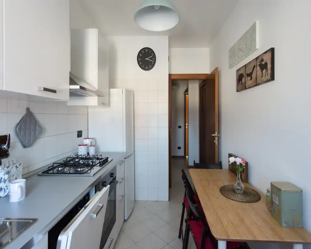Cucina lineare luminosa e ben attrezzata con piano a gas, forno, frigorifero e tavolo in legno per due. Pensili bianchi e decorazioni semplici creano uno spazio pratico e accogliente.