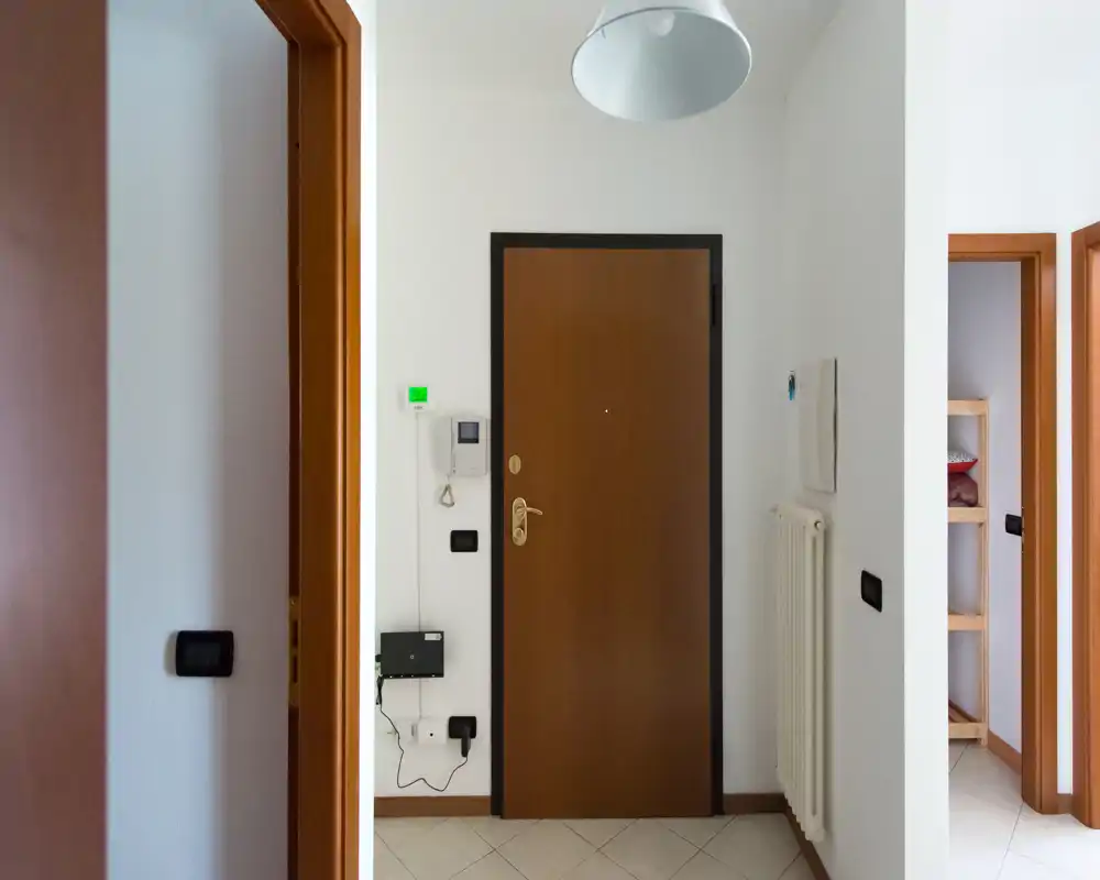 Ingresso compatto e ben illuminato con porta d'ingresso in legno, citofono, termosifone e pavimento piastrellato — inquadratura chiara e neutra adatta per l'annuncio.