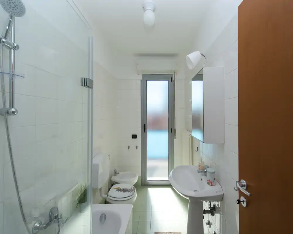 Bagno luminoso e moderno con vasca/doccia, lavabo a parete, bidet e WC; porta in vetro satinato che garantisce luce naturale e privacy.