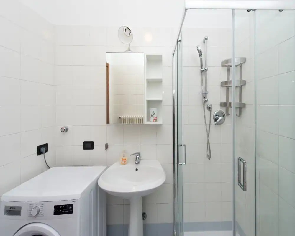 Bagno moderno e pulito con box doccia in vetro, lavabo a colonna e lavatrice; piastrelle bianche luminose e mensole semplici creano uno spazio ordinato e funzionale.