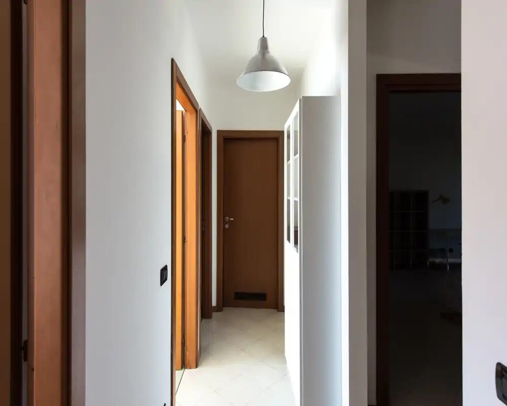 Ingresso luminoso e corridoio stretto con porte in legno, pavimento piastrellato e lampada a sospensione centrale — spazio d’ingresso pulito e ben illuminato.