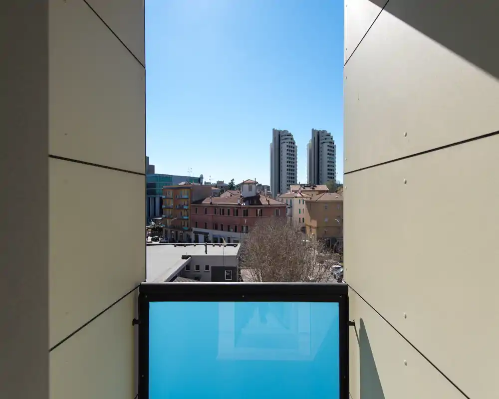 Piccolo balcone privato con pannello in vetro satinato blu, vista incorniciata sui tetti e cielo limpido — spazio esterno luminoso e tranquillo.